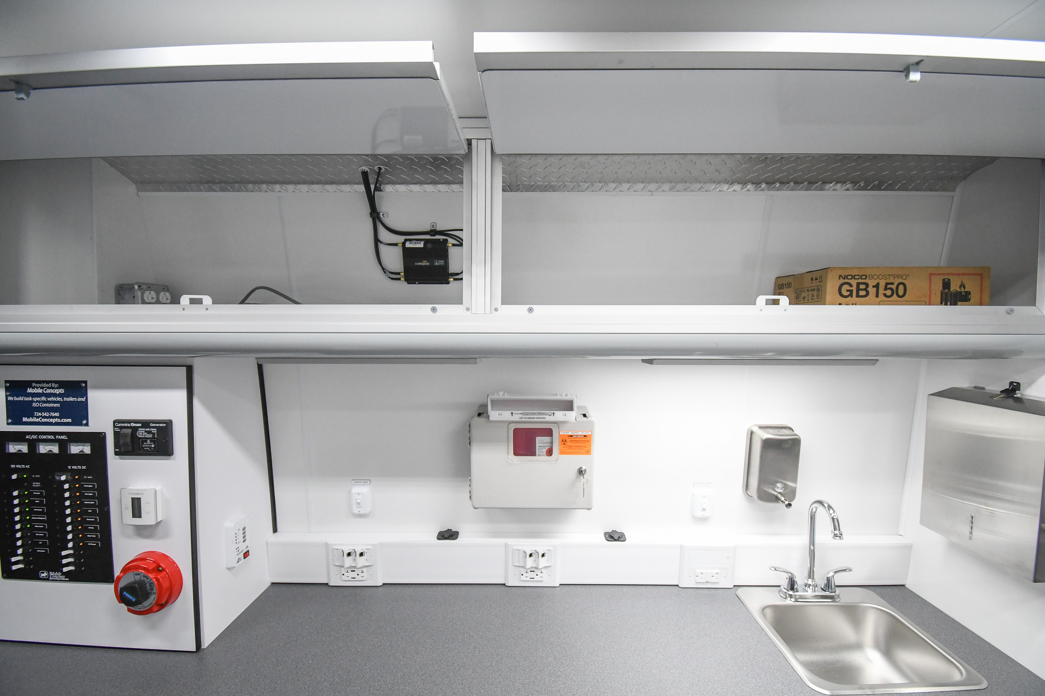 An alternate view of the cabinets, sink, and sharps disposal container inside the unit for Lancaster, PA. The overhead cabinet doors are open, allowing the Cradlepoint WiFi router to be seen.